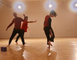 Three performers cluster towards the middle of the frame, in various blurred states of movement: arms reaching, legs akimbo, gaze down. Towards the bottom left hand corner is a small yoga block. The picture is brightly lit from sun-spotted lights in the back.