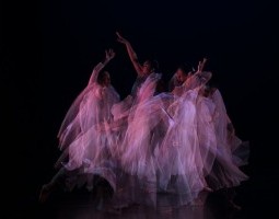 three dancers with large white blankets of tulle fabric pull them across each other, the camera catches them in a blurry flurry of motion. each dancer looks toward a different direction, arms to the sky. the photograph is a wash of the transparent tulle.