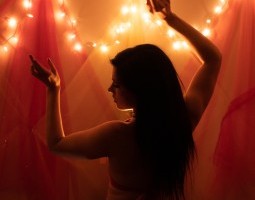 Yellow lights and gauzes of yellows and pinks set the stage. Seen from behind in silhouette, a belly dancer looks over her left shoulder and down. Her hands are raised slightly above her head.
