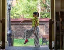 The image shows the dancer Eun Jung Choi in a green shirt and striped pants balancing on one leg with her other leg bent and raised. She is framed by an open doorway with the outdoor setting of the Arch Street Meeting House visible behind her, including a bench and brick wall, suggesting a serene environment for the dance.