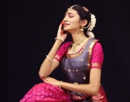 A woman in bright purple and pink short sleeves with gold trim, kneels, her left knee on the floor, the other obscured by the pleats of fabric between the legs of her costume. She rests her right hand, palm drawn with henna, gently against her cheek, seeming to hold that arm up with her left hand. Her face is tilted up, her eyes closed, and her mouth drawn tight as though pleading or sorrowful.