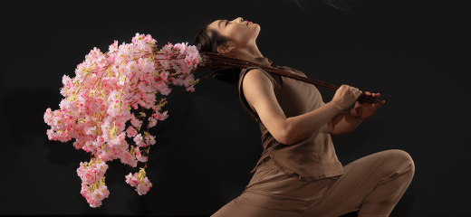 Weiwei Ma, dressed in a linen outfit, strikes a deep lunge against a dark background. With a branch of cherry blossom resting on her right shoulder, she arches her chest backward, gazing up toward the sky.