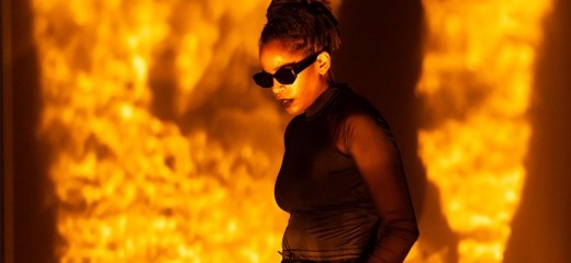 Angie Pittman stands with her hands in her pockets looking downward. She wears black sunglasses with her locs tied into a high ponytail. Pittman is doused in a textured golden amber light.