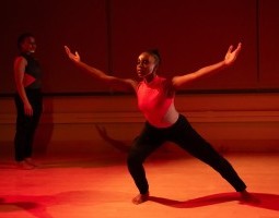 A dancer stands in a wide lunge with arms reaching wide and palms facing upward as if she is scooping the air above her. She wears a geometric red and grey leotard with black pants and a line of white face paint framing her chin. She is doused in a deep red light.