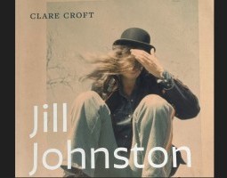 A woman in a black leather coat, jeans and a black bowler hat squats down. Her long hair is blown across her face obscuring it and her hand moves up to brush it away. The name Clare Croft appears in black letters in the top left corner. In large white letters, the bottom of the photo reads: Jill Johnston.