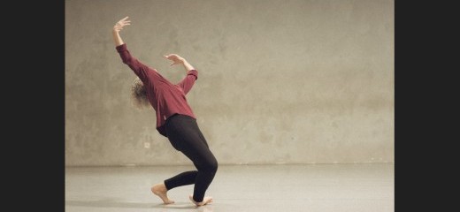 Positioned slightly to the left of center of the image, Lisa Kraus falls to the side over one ankle. Leaning back onto the ball of her other foot with her arms raised upward, her head lifts back, and her hair flies behind her. She is in a gray space wearing black leggings and a maroon long sleeve shirt. The photo is from 2025 and was inspired by the black and white photo by Lois Greenfield, described below under the heading “Homepage Image Description.”