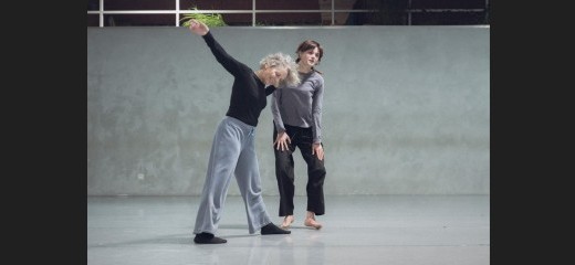 Smiling with her left foot stepped forward and right arm lifted, Lisa Kraus angles her weight lightly toward Laure de Dietrich. Kaus’s left hand rests on de Dietrich’s right arm. Both dancers have their heads inclined slightly to the left. de Dietrich may be descending from a small jump – both of her hands rest on her thighs, fingers spread, palms face-down, knees with a micro bend and feet supinated just off the ground. The duet takes place at Tictac Art Centre.