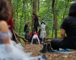 a dancer presses against the bare ground on all fours while another dancer stands barefoot on top of their back with the support of two others who hold her arms. They are wearing long-sleeved clothing in shades of grey, black and red. They are surrounded by trees and foliage. There are audience members in the foreground watching.