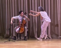 Tristan Price plays the cello seated on stage with a black microphone positioned in front of him. He wears black shoes, khaki pants and a white button-down shirt. He looks directly at dancer and his mother, Megan Bridge, who is dancing in the foreground, arching backward with one arm extending above her. Bridge wears a white top and grey cargo pants with sneakers.