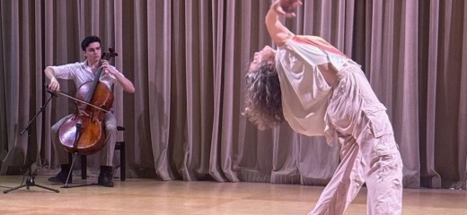 Tristan Price plays the cello seated on stage with a black microphone positioned in front of him. He wears black shoes, khaki pants and a white button-down shirt. He looks directly at dancer and his mother, Megan Bridge, who is dancing next to him with arms outstretched in his direction. Bridge wears a white top and grey cargo pants with sneakers.