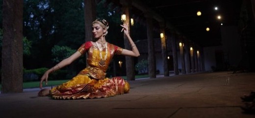 A woman sits on the stone floor of a long veranda at dusk, illuminated by hanging lanterns. The warm light makes her golden costume glow. Resting on her left hip, her right leg extends behind her, half bent. She looks down her extended right arm, letting her fingers drop toward the floor, while her left hand, at head level, pinches some fingers into her palm, leaving her ring and pinky fingers reaching up.