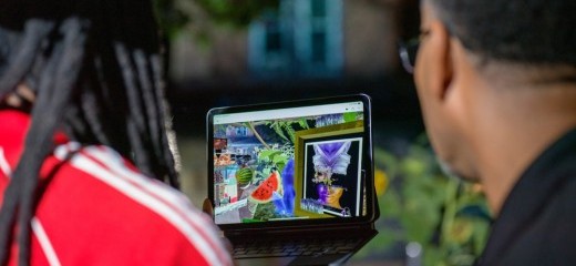 Two people look at an ipad with virtual reality images. Their backs are facing the camera and the person on the left wears a green hat, long locs, and a red shirt. The person on the right wears a black shirt and glasses.