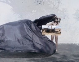 A composite still from Pajarillo, Como No Voy A Llorar? (Little Birdy, How Could I Not Cry). Travieso stands naked on concrete in front of an abstract gray digital background. With eyes closed, she holds a large black satin fabric that billows out to her left. Covering her body is an image of a large bird-like creature fallen on the ground in a desert landscape, with a child and adult looking on.