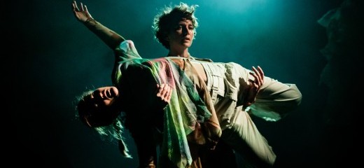 One woman, Zoe Rabinowitz, with green hued lighting behind her, is carrying horizontally across the front of her torso, Mary McGrath, who is lit with green and red hues across her outstretched body. Rabinowitz gazes forward beyond McGrath, who lies motionless on her back across the supporting arms of Rabinowitz.