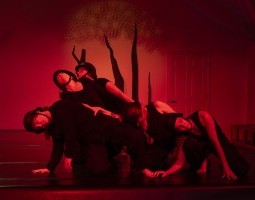 A group of dancers wearing all black with old-fashioned wartime helmets pile over one another, leaning onto each other with eyes tensely closed. A red light encapsulates the space, as large black spikes climb upwards along the back wall.