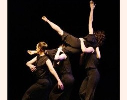 One dancer is elevated in a supported position while others provide grounded bases for a lift, demonstrating coordinated weight-sharing and balance. The movement highlights contemporary partnering, spatial precision, and controlled transitions.