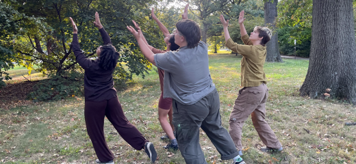 a group of four individuals stand together with arms lifted overhead making a clapping gesture with their hands. They are outside in the grass, surrounded by trees and other greenery. Their outfits are earth tones - browns, greens, and shades of grey.