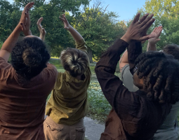 a group of four individuals stand together with arms lifted overhead making a clapping gesture with their hands. They are outside in the grass, surrounded by trees and other greenery. Their outfits are earth tones - browns, greens, and shades of grey.