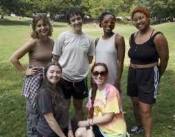Against a grassy, hilly field, six humans are collected in space. Four are standing in a line, two white individuals and two Black individuals, and two white individuals are kneeling facing each other in front of the standing people. They wear an assortment of loose and tight clothing and are all smiling with joy.