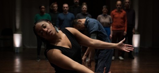 Shavon Norris stands in front of an audience wearing black clothing, large circular earrings, and a few finger rings. She is tilted to one side, facing the camera with eyes closed, arms outstretched in front of her, and palms facing upward.