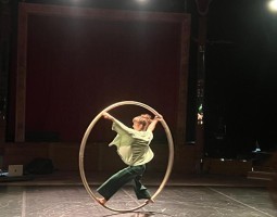 Hanschitz, a white woman wearing dark green pants and a flowy light green shirt, balances inside a human sized metal hoop, a Cyr wheel. Her arms and legs connect to the wheel in an X shape. The image is slightly blurry as if captured mid-motion.