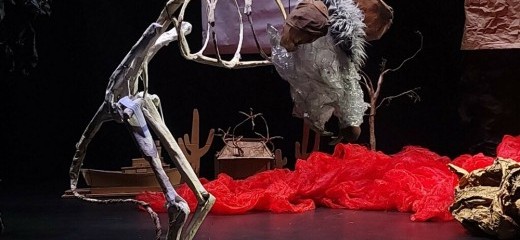 A large sculpture of a muskrat stands in a blackbox theatre. The muskrat is mostly skeleton with a furry head and ears. There is some red fabric on the floor in the background and a ball of red wiring sitting beneath the muskrat.