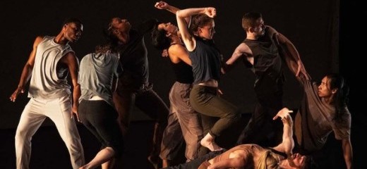 The image captures a dynamic moment from The Quake that Held Them All by Kayla Farrish. A group of dancers is intricately arranged, creating a sense of interconnectedness and tension. The performers are dressed in earthy, muted tones, and their movements range from grounded and angular to fluid and expansive. The dimly lit stage emphasizes their forms and the emotional intensity of the choreography, which explores themes of connection, resilience, and collective experience.