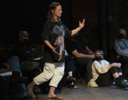 A woman is standing profile, mid-step, in a black shirt and white pants. Her arms are outstretched, palms face up. In the background audience members are seated on chairs and on the ground.