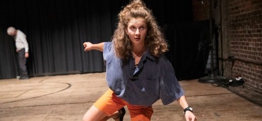 A dancer in a blue denim button down shirt and orange shorts is on one knee with her other leg raised and bent slightly behind her. She has long curly hair and peers out into the audience with her arms outstretched.