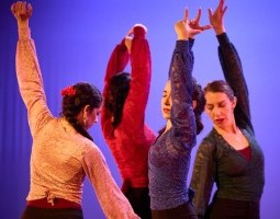 Four women stand in a group with their arms upraised and their heads in profile. They are wearing long-sleeved shirts in yellow, red, navy blue, and black.