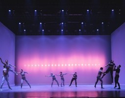 In groups of three, dancers are positioned stage left, stage right and centerstage. The left and right groups perform a lift with two dancers at the base and the aerial dancer reaching outward. Through reaching in similar form to the aerial dancers, the centerstage trio remains grounded. Dancers stand against a white backdrop with pink and blue lights. They wear uniform spandex pants and crop tops.