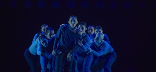 In blue lighting, a crowd of dancers hover over one another looking forward into the camera lens. One dancer stands in the forwardmost position with the group grasping their shoulders, arms, and legs, as if to constrain their forward motion.