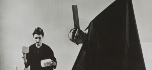 A black and white photo of Lucia Dlugoszewski kneeling on the floor holding a cylinder and a box shaped item in either hand. in front of her sits a pile of wooden objects and instruments. beside her, Hawkins, draped in a black cape-like garment which reveals only his head, tilts toward Dlugoszewski and creates a triangular shape with the garment.
