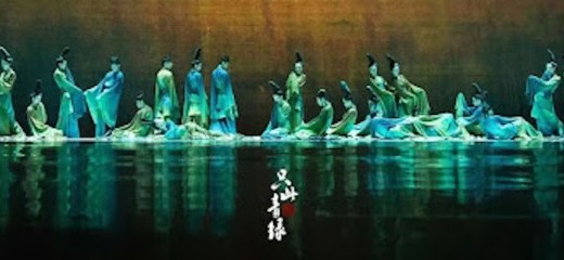 This image showcases a breathtaking tableau from The Journey of a Legendary Landscape Painting. A large ensemble of dancers, clad in flowing blue and green costumes, is positioned across the stage in dynamic, yet harmonious formations. Their fluid movements and graceful postures mirror the essence of a traditional Chinese landscape, with cascading lines that evoke rivers and mountains. The reflective stage floor enhances the ethereal atmosphere, creating a mirror-like effect that doubles the dancers’ artistry and immerses the audience in a dreamlike scene.
