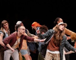A group of standing performers lean towards one another while gazing intently towards an unknown subject. One performer, wearing a maroon jacket and glasses, grips the body of the jacket of a performer with long hair wearing a baseball cap. Their facial expressions evoke intensity, but their bodies appear to be frozen in time.