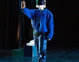 white person wears an astronaut helmet with the face shield down, though it doesn’t quite cover their chin. They stand with one foot in front of the other and hold up an arm, softly opening their palm to face forward as if waving or saying “hold up”. Turquoise light makes shapes on their open palm and their neck. They’re dressed in textures of blue, glimmering pants and a blue bomber with cinched ruffled seams along the arms. Two stage blocks rest behind them, one with a pair of plush white boots set atop.
