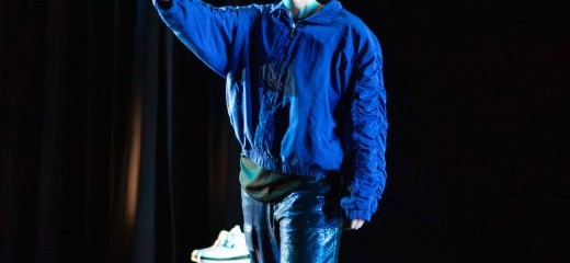 white person wears an astronaut helmet with the face shield down, though it doesn’t quite cover their chin. They stand with one foot in front of the other and hold up an arm, softly opening their palm to face forward as if waving or saying “hold up”. Turquoise light makes shapes on their open palm and their neck. They’re dressed in textures of blue, glimmering pants and a blue bomber with cinched ruffled seams along the arms. Two stage blocks rest behind them, one with a pair of plush white boots set atop.
