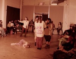 A warm, slightly grainy, tinted photo of an in-the-round white-walled performance space. At the center of the room a performer in polka-dot pants lies on the ground. Above her, three performers gather, one wearing a shirt that says “Join the Hike.” To their left, Corinne Jones – wearing a white button-up and brown tights – gestures out to the audience with an open arm.