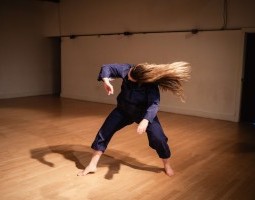 Kay MacMaster wearing blue coveralls stands in the center of a wood paneled performance space. Their body is folded forward, knees slightly bent, and the head tossed sideways with their long blonde hair dramatically fanning out, capturing a feeling of wild abandon and emotional release.