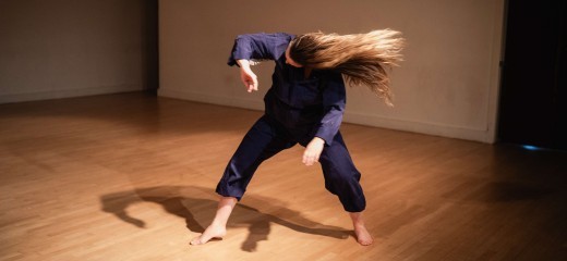 Kay MacMaster wearing blue coveralls stands in the center of a wood paneled performance space. Their body is folded forward, knees slightly bent, and the head tossed sideways with their long blonde hair dramatically fanning out, capturing a feeling of wild abandon and emotional release.