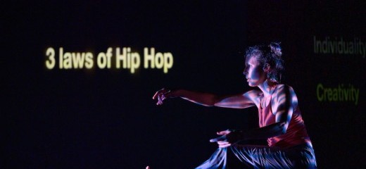 One dancer balances her bent right leg, left leg lifted, out stretched to match her opposite arm.Words to the left of the frame written on the back wall read “3 laws of hip hop.” The rest of the stage is nearly black.