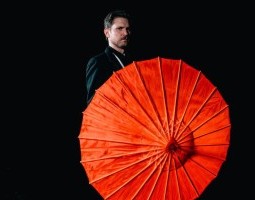 Against a black background, Schreiben wearing a black suit stands partially obscured by the vibrant red parasol he is holding.