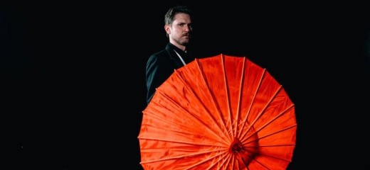 Against a black background, Schreiben wearing a black suit stands partially obscured by the vibrant red parasol he is holding.