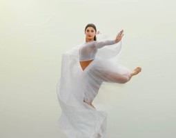 Abby Donnenfeld appears to have just landed an arabesque leap. She stands on one bent leg, enwrapped in billows of white tool, her other leg abducted, knee bent and toes pointing backward. Her left arm extends to the side, wrist flexed, she holds the opposite end of the billowing tool between her lengthened fingers. Her right arm, extending behind her head, isn’t visible. She looks definitely towards a spot just left of the camera, hair smoothed back, lips red, face relaxed and focused. The background is entirely white and nearly envelopes her white fabric.