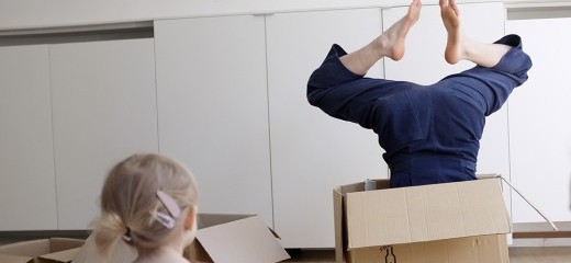 Selena Rook stands in a white-walled room wearing a sleeveless navy blue jumpsuit. Perched on her right hip is a medium sized cardboard box.