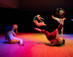 The ensemble erupts into explosive movement—some in deep lunges, others mid-air—surrounded by brilliant bursts of blue, yellow, orange, pink, and red light that flood the space.