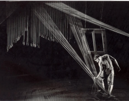 A black and white still of a scene from Daniel Stein’s Inclined to Agree, 1986. A muscular Stein lunges his bent left leg upstage, bending his torso to his left, wearing dress slacks , movement shoes and shirtless. He is stepping on two bunches of taught chords that run diagonally from his right ankle up to the rafters where more chords of varying lengths hang with weights attached . An empty door frame hangs from chords just upstage of Stein, it tilts towards him on a steep incline.