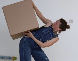 Two feet dangle in the air attached to a person wearing a blue jumpsuit. The person is upside down. Her head is not visible; it is inside a cardboard box. One child with blond pigtails stands to the left of the box looking on.