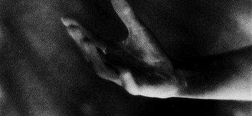 In greyscale photograph, a hand emerges from the dark, fingers shades of white and grey like brushstrokes.