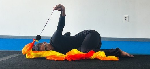 Performer Daisie Cardona rests in a fetal position on her side with her eyes closed. Dressed all in black, one arm stretches above her body, holding a black string taut. The string is connected to a red, orange, and yellow ribbon which rests on the ground.